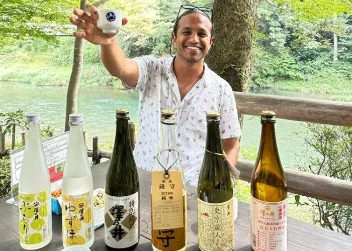 A man raising a cup of sake to the camera while smiling. In front of them, many bottles of sake.